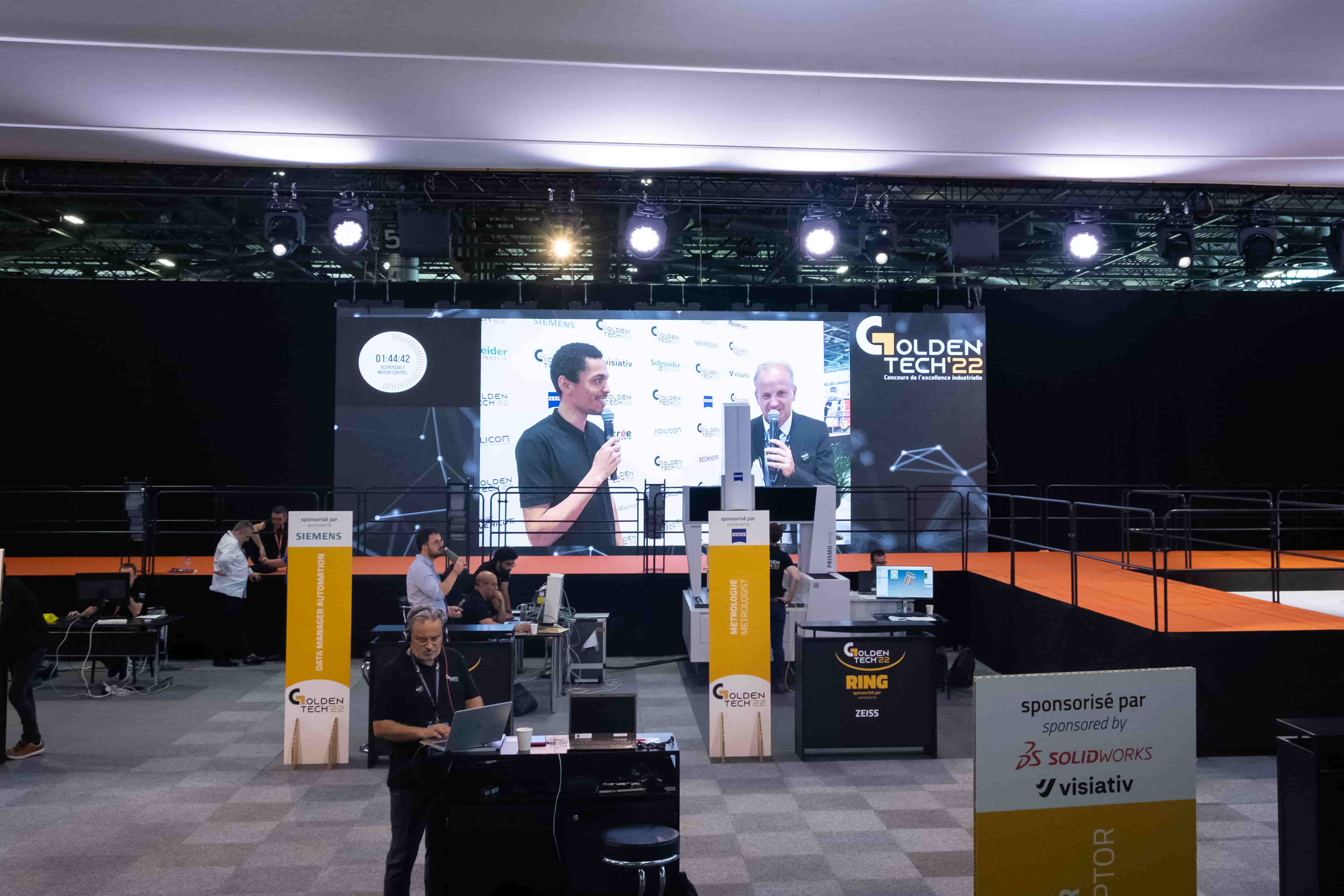 Stand Golden Tech avec un écran géant en fond où est diffusée l'interview d'un participant au concours par le présentateur.