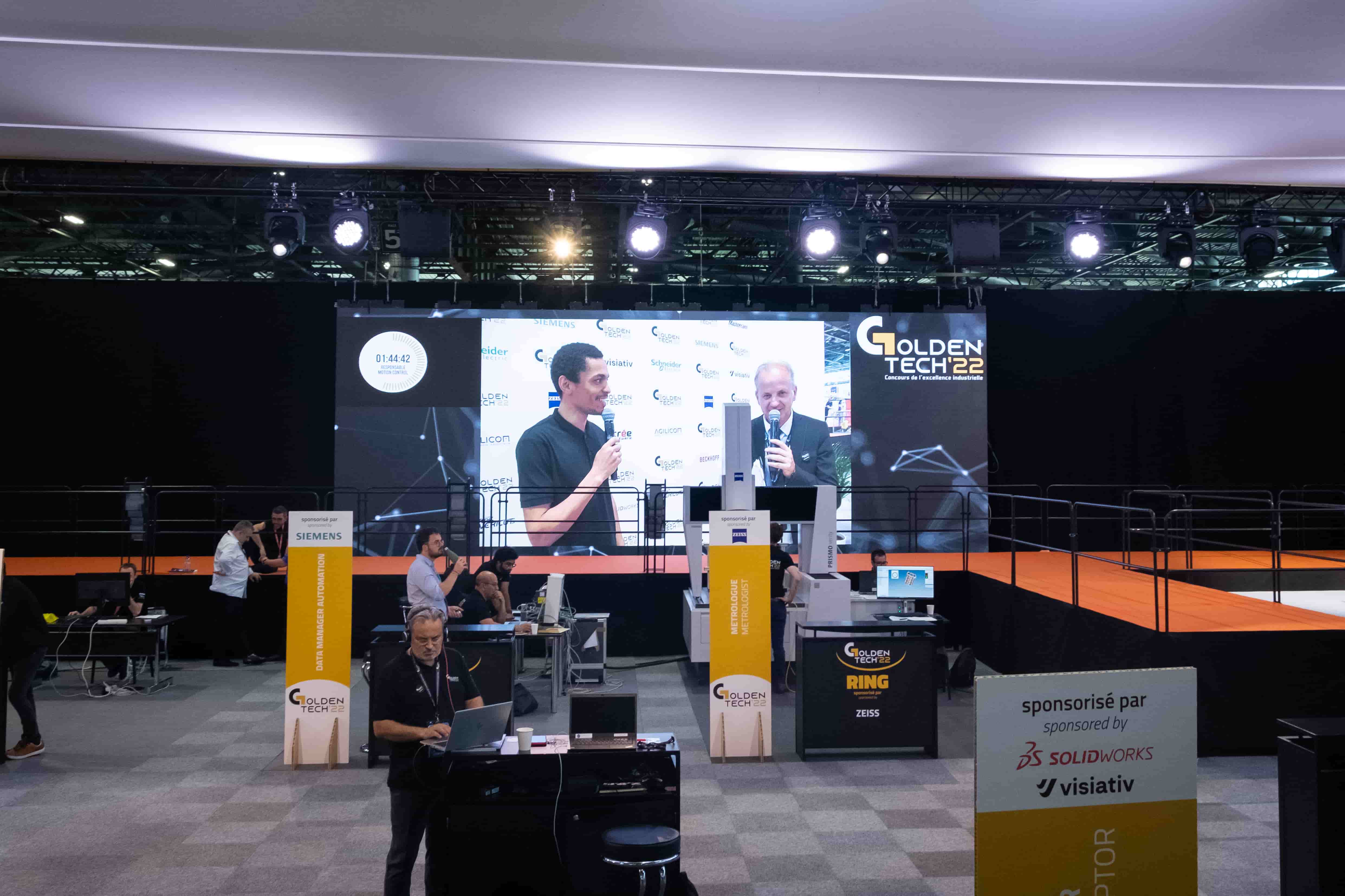 Stand Golden Tech avec au fond un écran géant où est diffusée une interview d'un participant au concours par le présentateur.